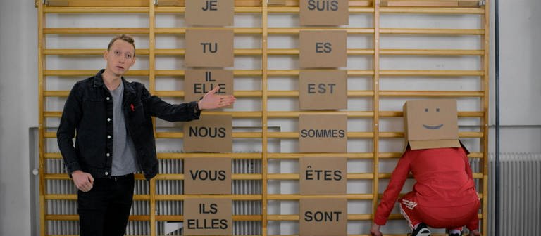 Ein junger Mann vor einer Sprossenwand, an ihr hängen Schilder. Auf den Schildern stehen französische Pronomen und die Konjugationen von "Être". Daneben sitzt eine Person mit Karton auf dem Kopf. (Foto: WDR)