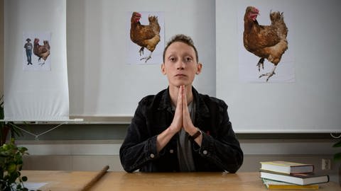 Ein junger Mann sitzt mit gefalteten Händen an einem Tisch, hinter ihm verschiedene Bilder von einem Huhn. (Foto: WDR)