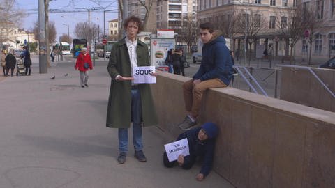 Ein junger Mann steht in einer Stadt und hält ein Schild, darauf steht "sous". Daneben kauert ein Junge unter einem Jungen auf einer Mauer und hält ein Schild mit "un monsieur".  (Foto: WDR)