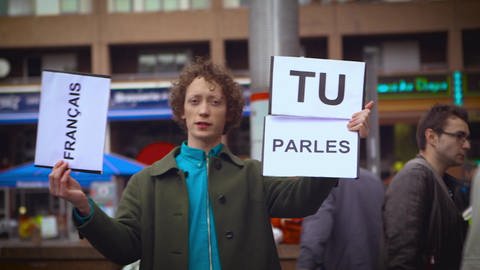 Ein junger Mann steht auf der Straße und hält Schilder hoch, darauf steht "Tu parles français" (Foto: WDR)