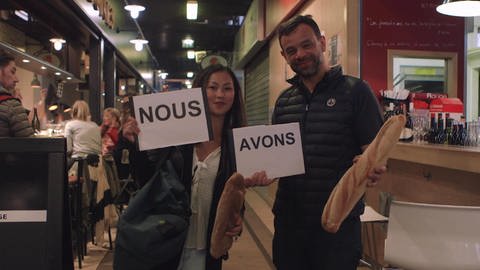 Eine Frau und ein Mann stehen in einem Restaurant. Sie halten Schilder hoch mit "Nous" und "avons", der Mann hält ein Baguette. (Foto: WDR)