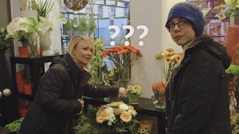 Ein Junge und eine Verkäuferin stehen in einem Blumenladen, neben seinem Kopf schweben drei Fragezeichen. (Foto: WDR)