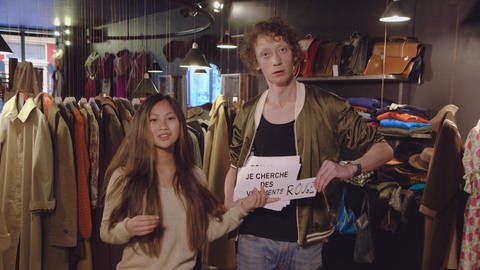 Ein junger Mann und eine junge Frau stehen in einem Kleiderladen. Der Mann hält ein Schild mit der Aufschrift "Je cherche des vêtements rouge". (Foto: WDR)