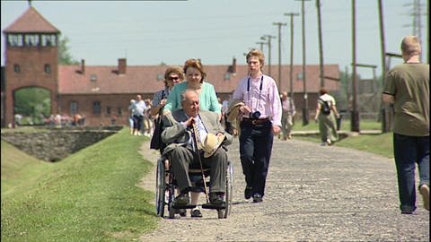 Menschen laufen durch das Konzentrationslager Auschwitz-Birkenau. Eine Frau schiebt einen älteren Mann im Rollstuhl. (Foto: WDR)