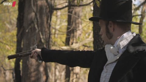 Ein Filmausschnitt von einem Mann in Wrack und Zylinder, der im Wald steht und mit einer Pistole zielt. (Foto: WDR)