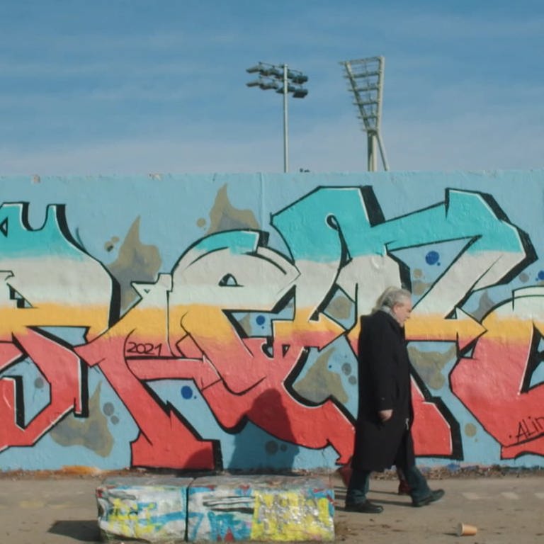 Zwei Menschen laufen im heutigen Berlin vor der Mauer, die mit Graffiti bespüht ist. Die Teilung Deutschlands hat ihr Leben geprägt. (Foto: SWR)