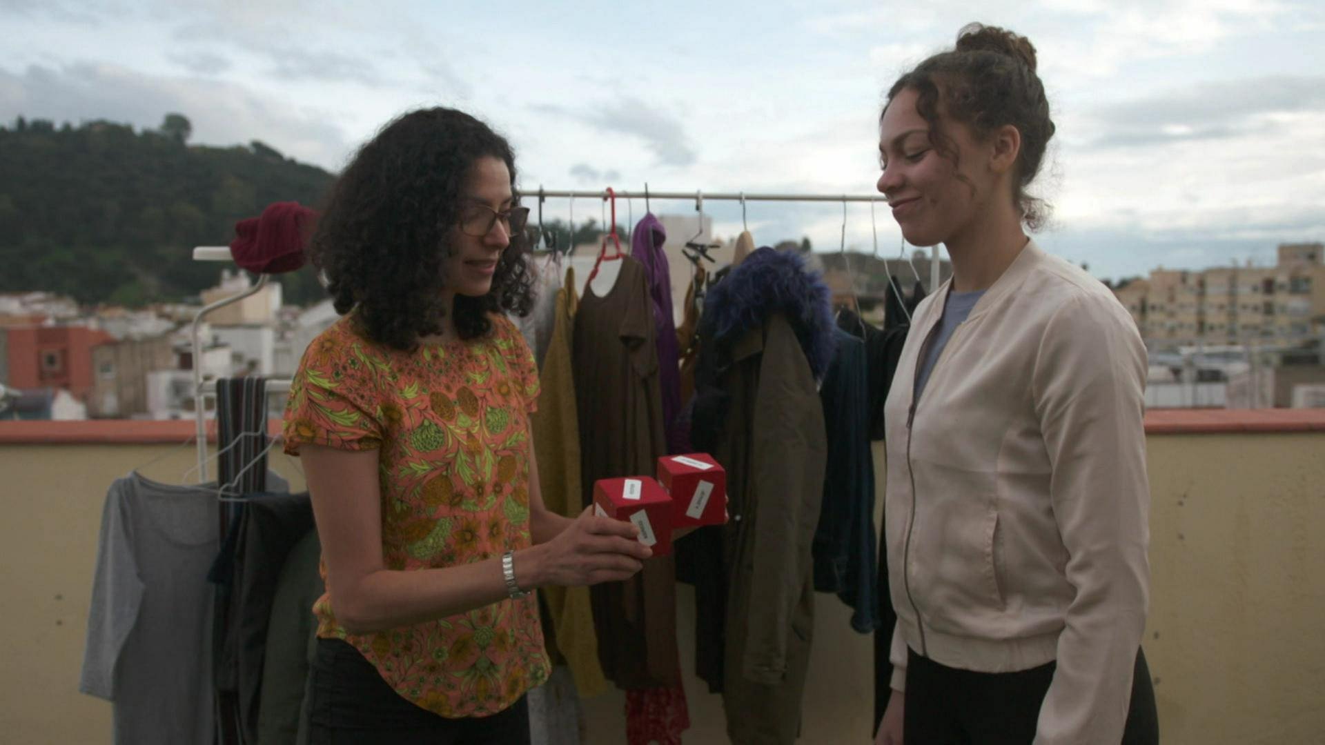 Zwei Frauen stehen auf einer Terasse vor einer Kleiderstange. Die eine Frau hält der anderen zwei rote Würfel entgegen. (Foto: WDR)