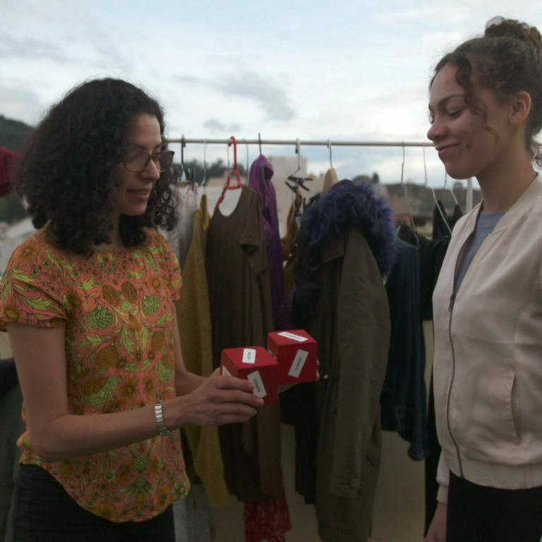 Zwei Frauen stehen auf einer Terasse vor einer Kleiderstange. Die eine Frau hält der anderen zwei rote Würfel entgegen. (Foto: WDR)