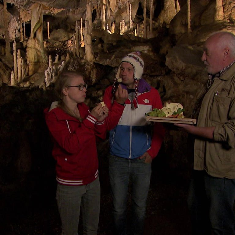 Ein Mann und eine Frau stehen in einer Tropfsteinhöhle und essen Käse. Ein älterer Mann hält ihnen eine Käseplatte hin.  (Foto: WDR - Screenshot aus der Sendung)