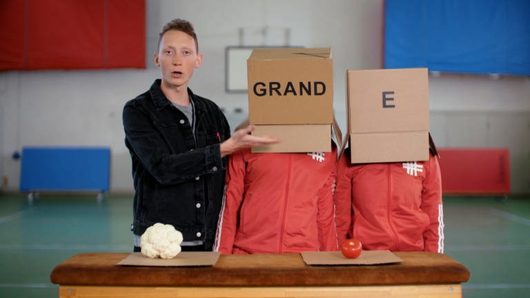Ein junger Mann steht in einer Turnhalle, neben ihm zwei Personen mit Kartons auf dem Kopf. Auf einem Karton steht "Grand", auf dem anderen "e". (Foto: WDR)