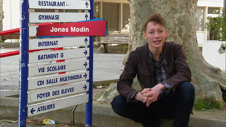 Ein junger Mann sitzt vor einem französischen Wegweiser. (Foto: WDR - Screenshot aus der Sendung)