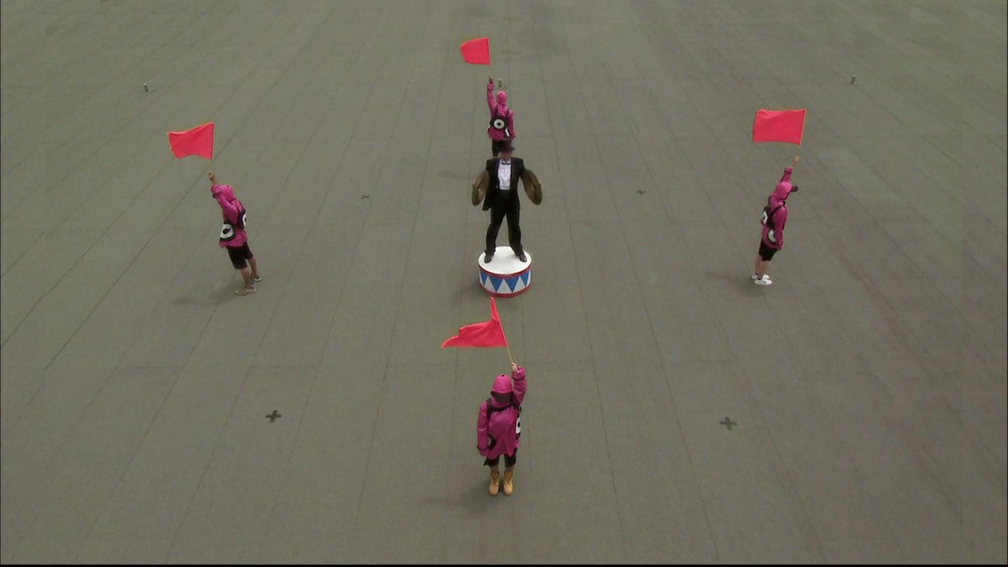 Eine schwarz gekleidete Person mit Zylinder und zwei Becken steht auf einem Platz, um ihn herum stehen vier Personen und halten Fahnen in die Luft. (Foto: SWR - Screenshot aus der Sendung)