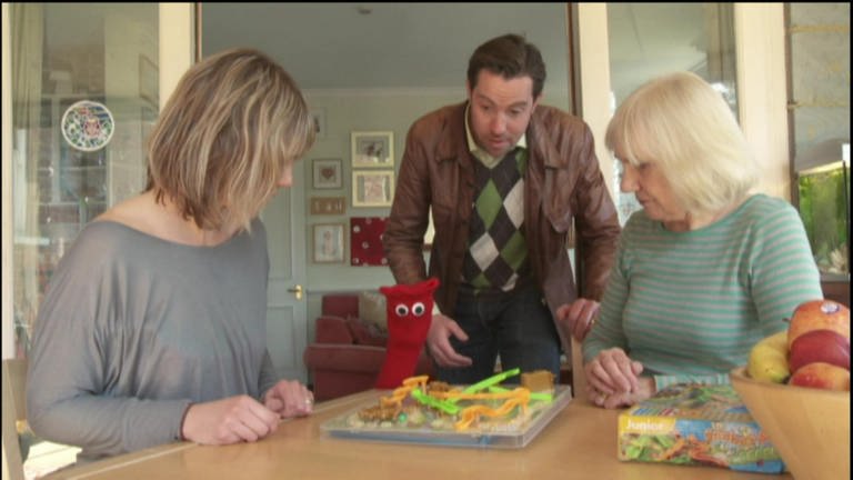Drei Personen und eine rote Strumpfhandpuppe sind an einem Tisch versammelt und spielen ein Spiel. (Foto: WDR - Screenshot aus der Sendung)