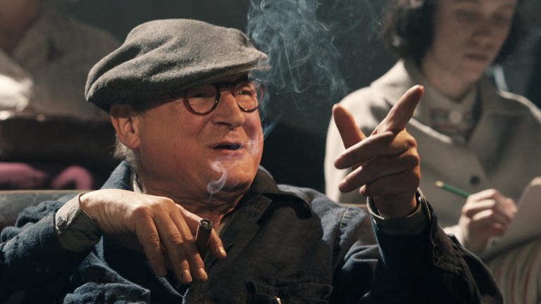 Ein Mann mit Hut und Brille raucht Zigarre und zeigt mit dem Finger auf etwas. (Foto: WDR)
