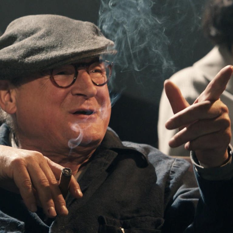 Ein Mann mit Hut und Brille raucht Zigarre und zeigt mit dem Finger auf etwas. (Foto: WDR)