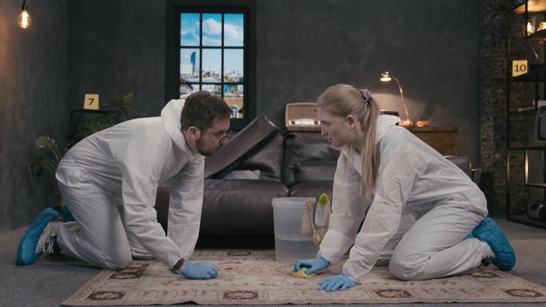 Zwei Personen in Schutzanzügen knien auf einem Teppich und reinigen ihn mit Schwämmen. (Foto: SWR)