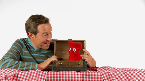 Ein Mann findet eine rote Strumpfhandpuppe in einer Holzkiste.
