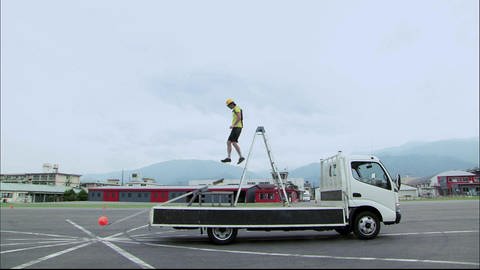 Eine Leiter steht auf einer Autoladefläche. Eine Person springt von dieser Leiter auf eine Wippe, auf der anderen Seite liegt ein Ball.