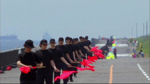 Schwarzgekleidete Personen stehen in einer Reihe und halten rote Fahnen.