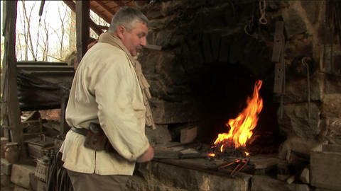 Ein Schmied steht an einem offenen Feuer und bearbeitet Werkzeug. 
