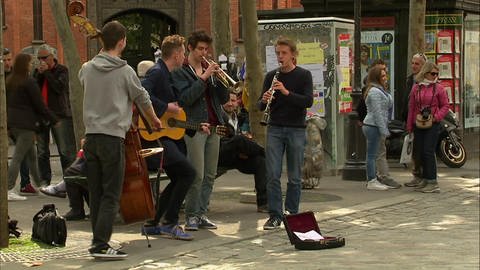 Junge Menschen stehen in einer Einkaufsstraße und machen Straßenmusik. 