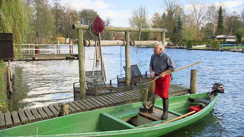 Ein Mann steht auf einem kleinen, grünen Boot. Er hält einen Kescher und trägt Gummistiefel und Schürze.
