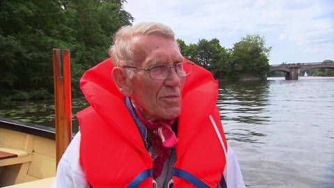 Ein älterer Mann steht mit einer roten Rettungsweste an einem Fluss.