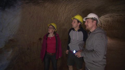 Drei Personen sind in einer Höhle und tragen Helme und eine Taschenlampe.