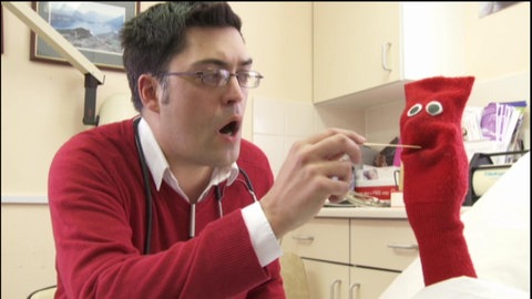 Ein Arzt hält einer roten Strumpfhandpuppe einen Holzstab in den geöffneten Mund.