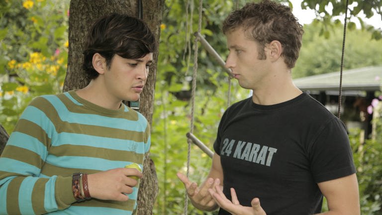 Zwei junge Männer stehen in einem Garten und unterhalten sich.