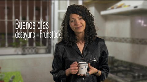 Eine Frau steht in Schlafanzug und mit Kaffetasse in einer Küche. Neben ihr der Schriftzug "Buenos dìas, desayuno = Frühstück"