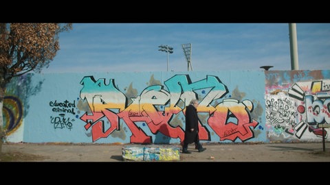 Zwei Menschen laufen im heutigen Berlin vor der Mauer, die mit Graffiti bespüht ist. Die Teilung Deutschlands hat ihr Leben geprägt.