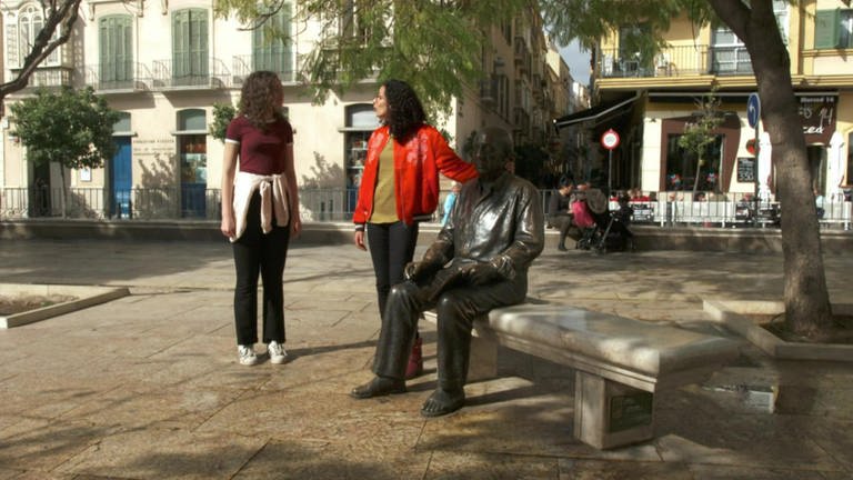 Zwei Frauen stehen auf einem Platz. Eine der beiden legt ihre Hand auf eine Bronzestatue eines Mannes, der auf einer Bank sitzt.