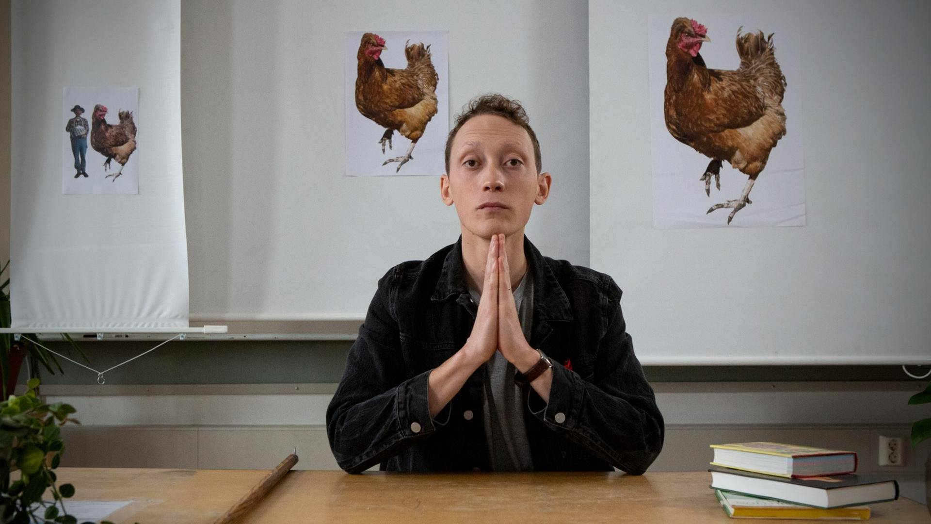 Ein junger Mann sitzt mit gefalteten Händen an einem Tisch, hinter ihm verschiedene Bilder von einem Huhn.