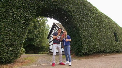 Ein Mann und eine Frau stehen vor einer sehr hohen Hecke und schauen auf einen Kompass.