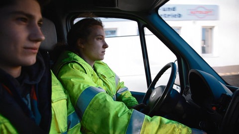 Eine junge Frau und ein junger Mann sitzen im Auto. Beide tragen neongelbe Arbeitsjacken.