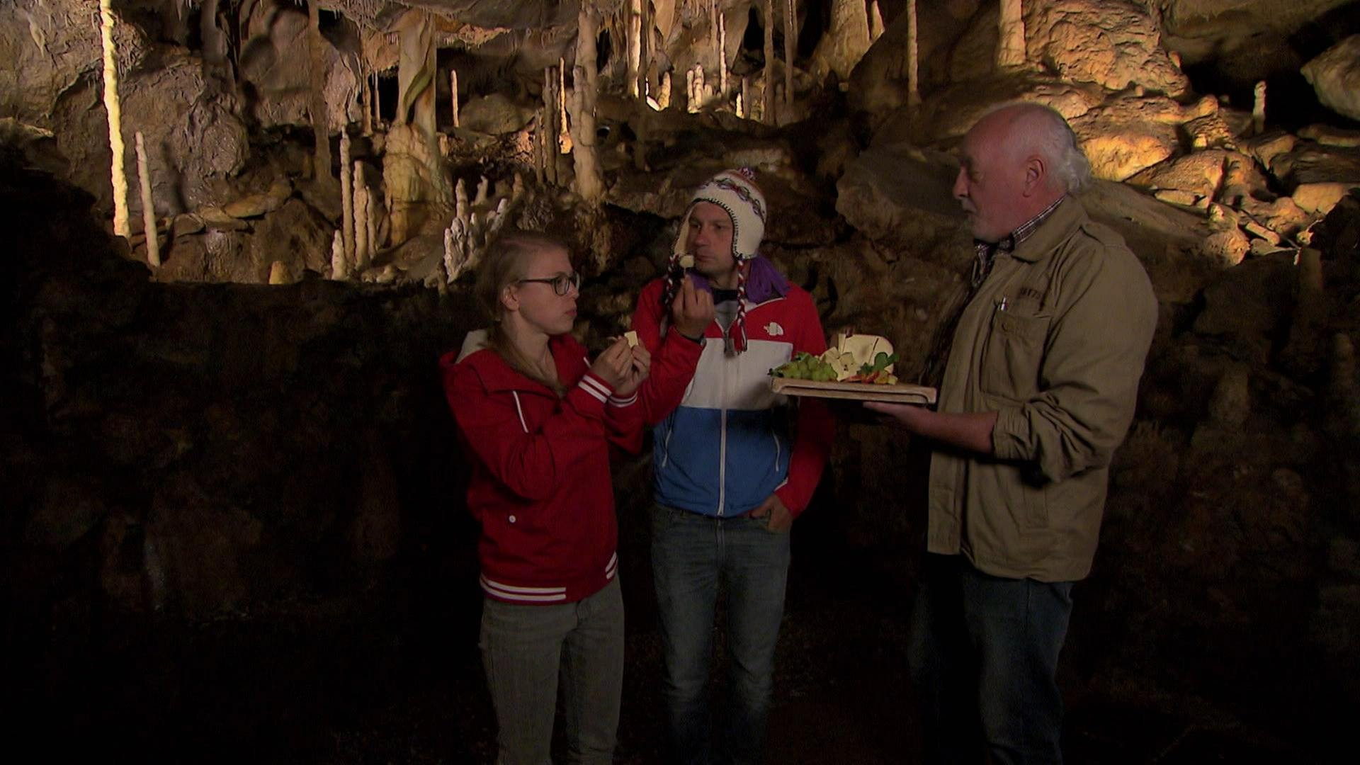 Ein Mann und eine Frau stehen in einer Tropfsteinhöhle und essen Käse. Ein älterer Mann hält ihnen eine Käseplatte hin. 
