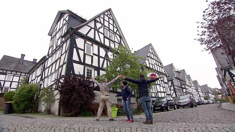 Drei Menschen stehen vor einem Fachwerkhaus. Zwei heben die Arme hoch, in der Mitte steht die dritte Person.