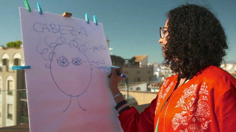 Eine Frau zeichnet einen Kopf an eine Tafel und schreibt die spanischen Namen der Körperteile auf.