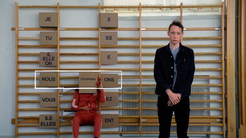 Ein junger Mann steht vor einer Sprossenwand, an ihr hängen Schilder. Darauf stehen französische Pronomen und die Endungen konjugierter Verben auf -er.