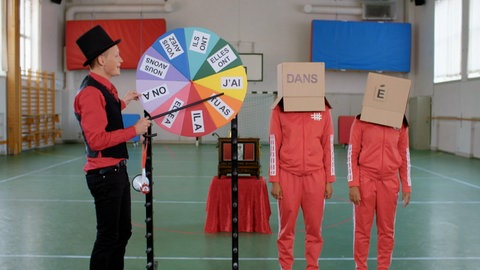 Ein junger Mann steht an einem bunten Glücksrad, hinter ihm zwei Personen mit Kartons auf dem Kopf. Auf einem Karton steht 