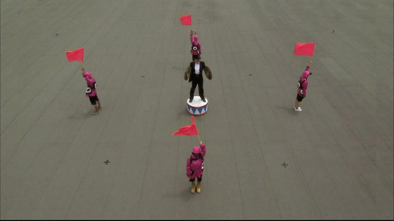 Eine schwarz gekleidete Person mit Zylinder und zwei Becken steht auf einem Platz, um ihn herum stehen vier Personen und halten Fahnen in die Luft.