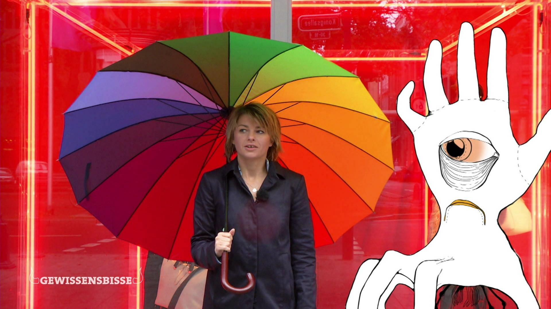 Die Moderatorin steht mit einem Regenschirm in Regenbogenfarben vor einem roten Schaufenster, neben ihr eine Zeichnung der Habgier als Hand.