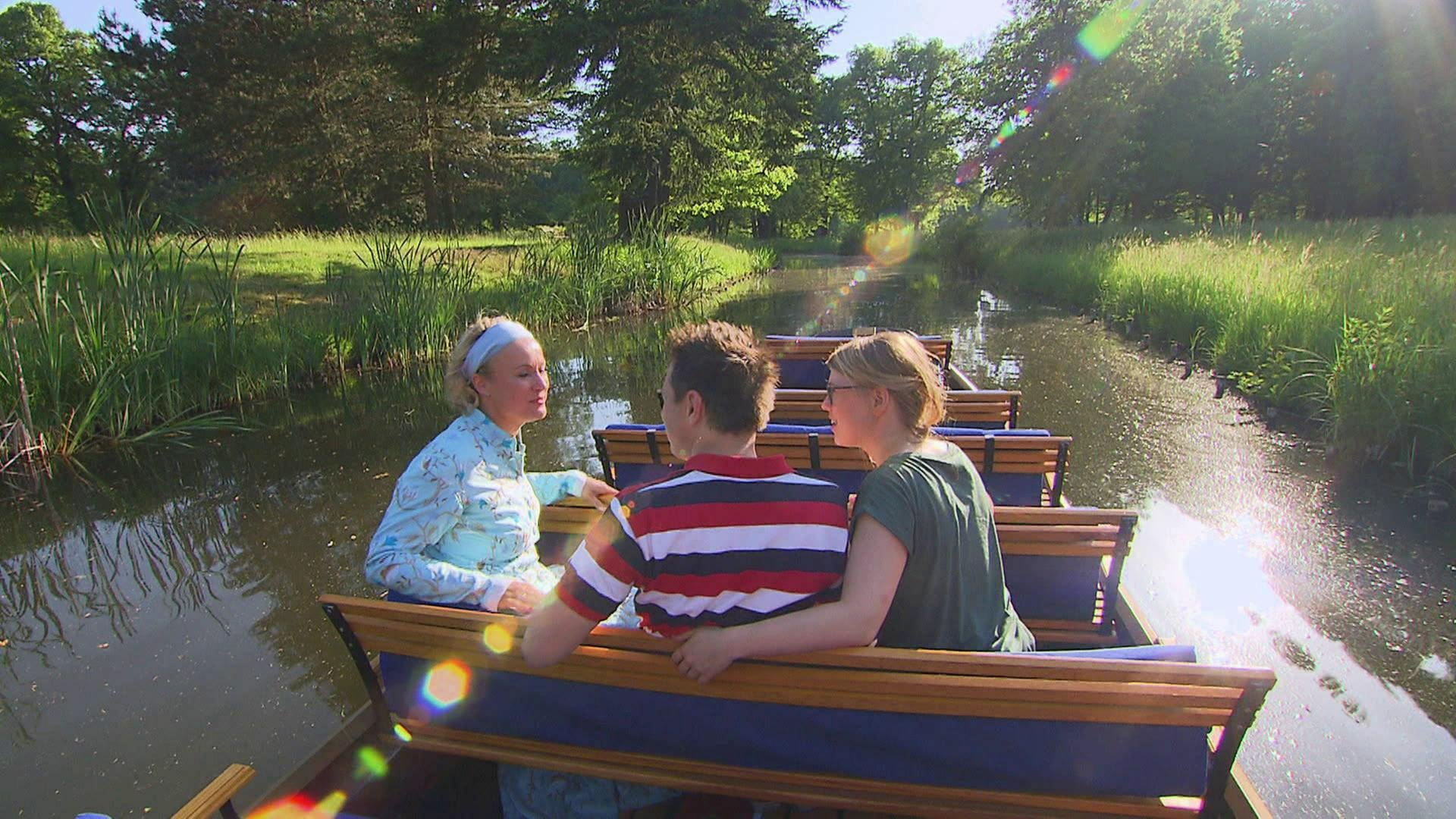 Drei Personen sitzen auf einem Boot und fahren über einen Kanal durchs Grüne. 