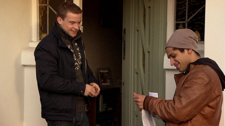 Zwei junge Männer stehen grinsend vor einer offenen Haustür.