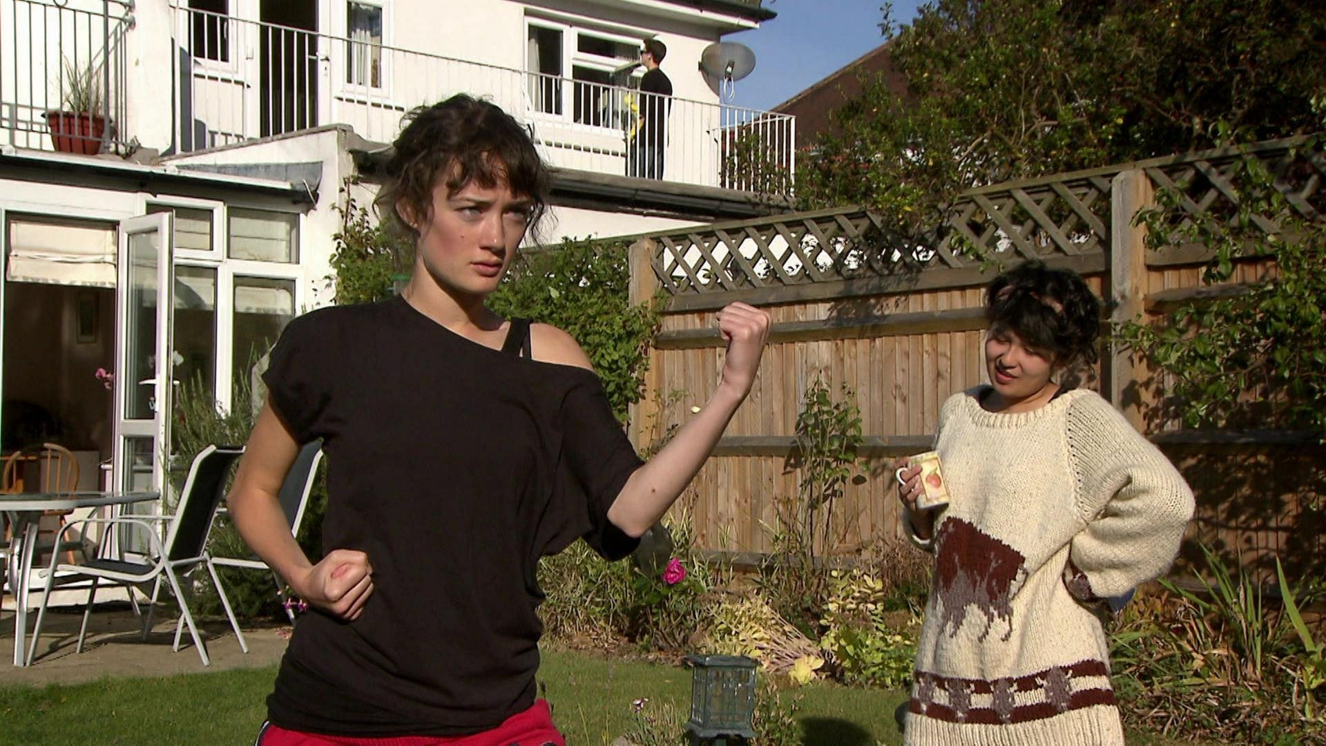 Zwei junge Frauen stehen im Garten. Die eine trinkt Kaffe, die andere trainiert Karate.