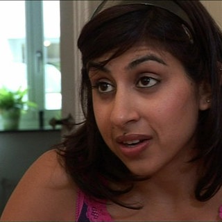 Eine junge Frau sitzt in einer Küche und spricht mit hochgezogenen Augenbrauen.