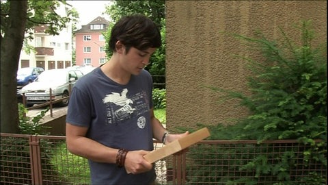 Ein junger Mann steht vor einem Haus und schaut traurig auf ein gerahmtes Foto in seinen Händen.