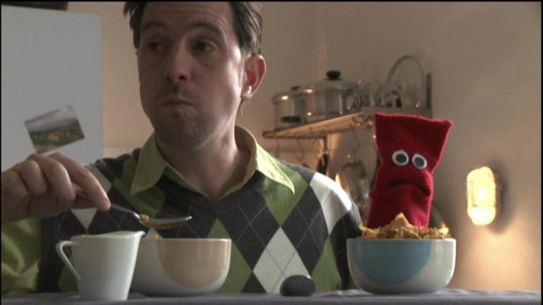 Ein Mann und eine rote Strumpfhandpuppe sitzen am Tisch und frühstücken.
