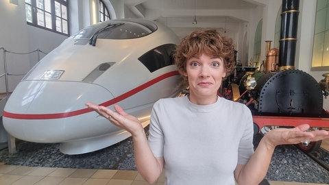 Moderatorin Lena Ganschow steht zwischen einem ICE und einer Dampflok